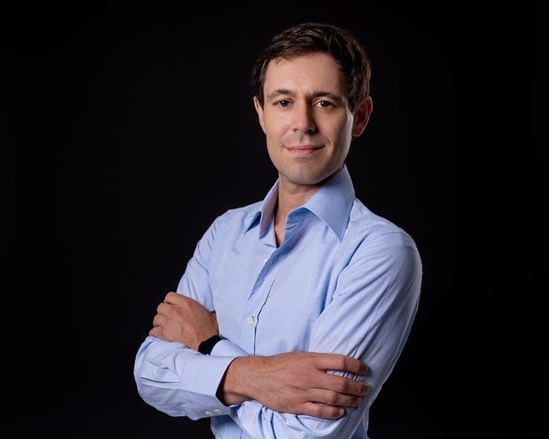 Professional headshot of John Arrow in a light blue dress shirt against a dark background