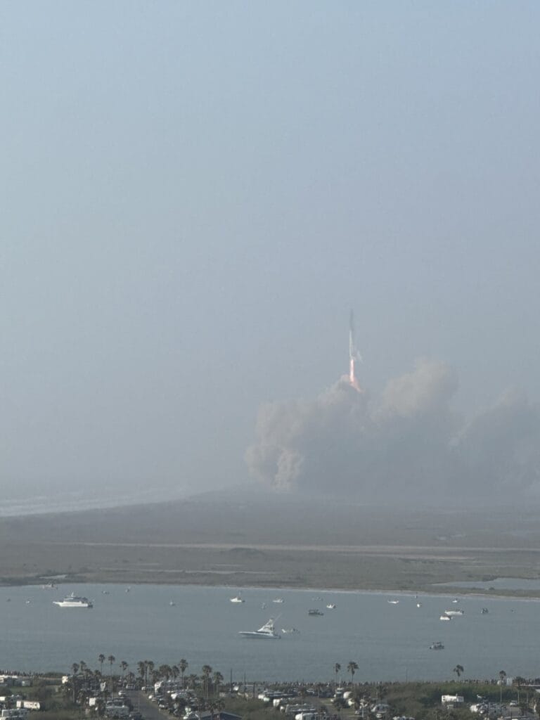 SpaceX Starship lifting off in the distance, viewed across a bay lined with boats and spectators